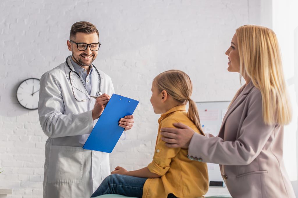 Uśmiechnięty lekarz z podkładką patrząc na dziewczynę w pobliżu rodzica na niewyraźnym pierwszym planie  - Zdjęcie, obraz