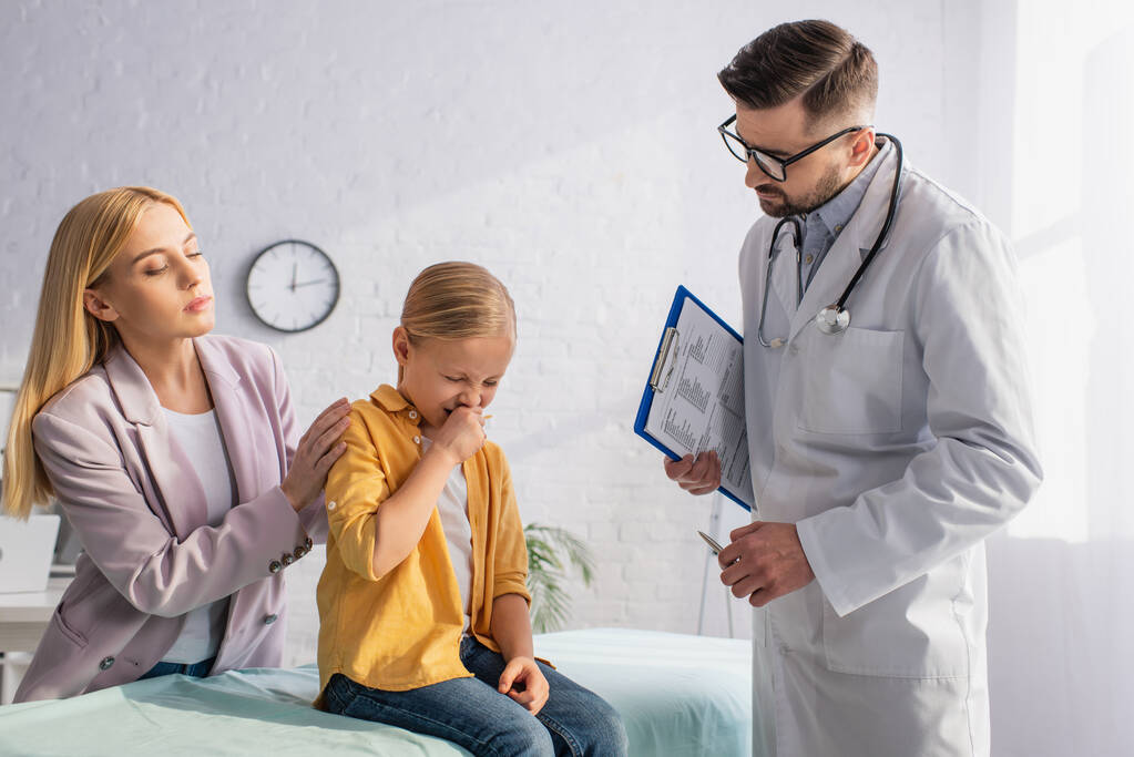 Sick kid sneezing near mother and family doctor with clipboard  - Photo, Image