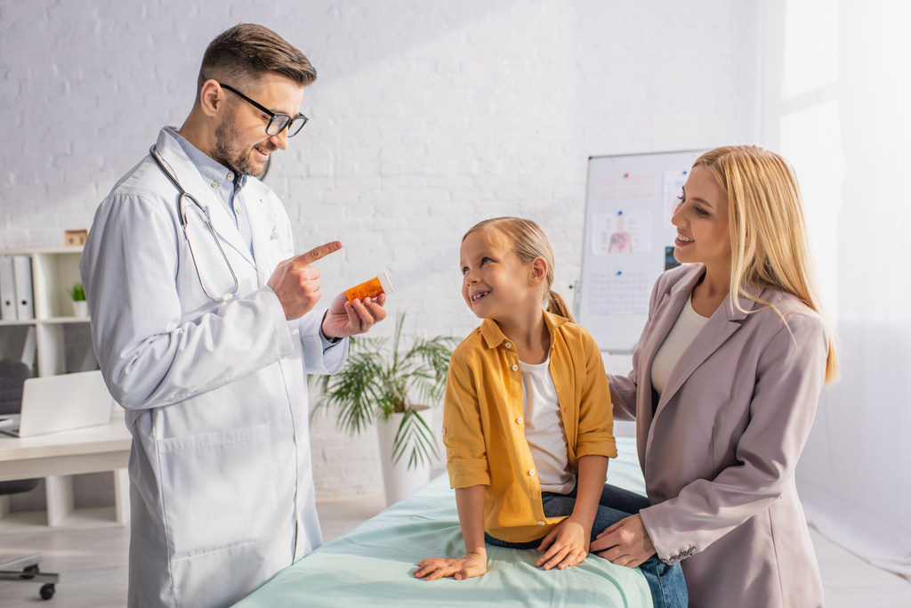 Medico sorridente che tiene le pillole e indica con il dito la madre e il bambino sul divano medico  - Foto, immagini