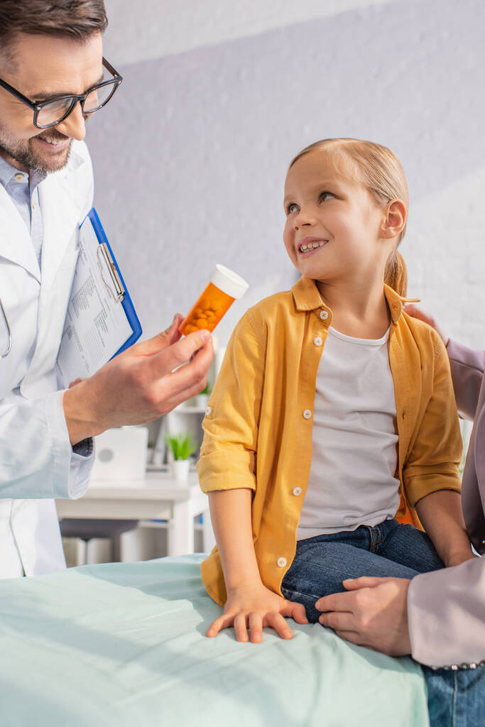 Позитивний лікар з буфером тримає таблетки біля усміхненої дівчини на медичному дивані і жінці
  - Фото, зображення