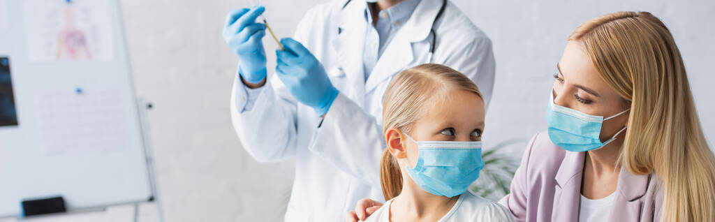 Мать и ребенок в медицинских масках рядом размытый семейный врач с вакциной, баннер  - Фото, изображение
