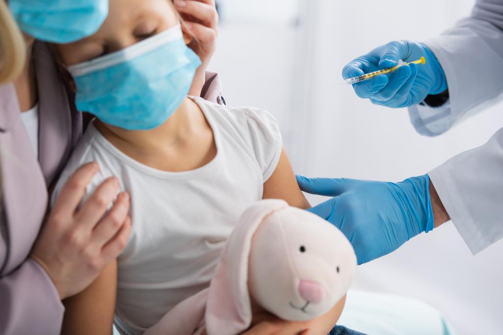 Доктор в латексных перчатках держит шприц рядом с матерью обнимающей ребенка в медицинской маске  - Фото, изображение