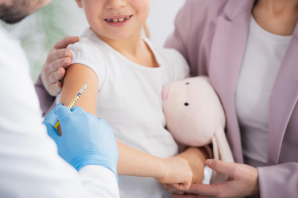 Cropped view of doctor holding syringe near smiling kid with soft toy and mother  - Photo, Image