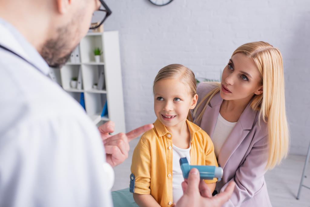 Frau schaut Hausarzt an und zeigt auf Inhalator neben lächelnder Tochter  - Foto, Bild