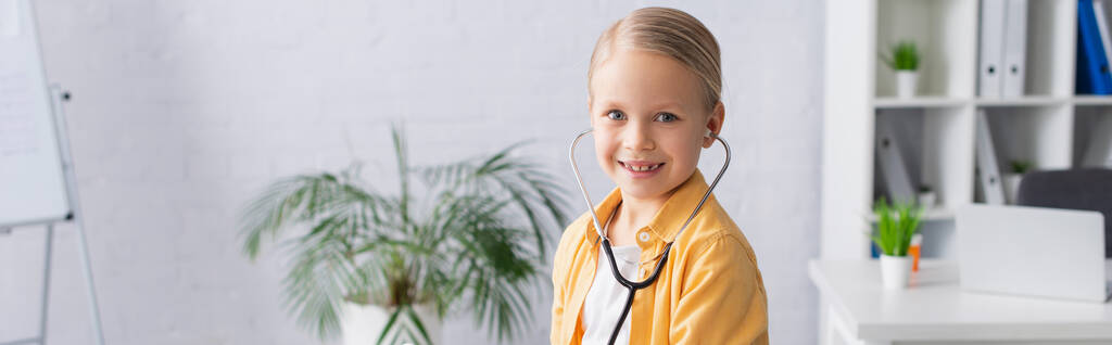 Позитивный ребенок в стетоскопе смотрит в камеру в клинике, баннер  - Фото, изображение