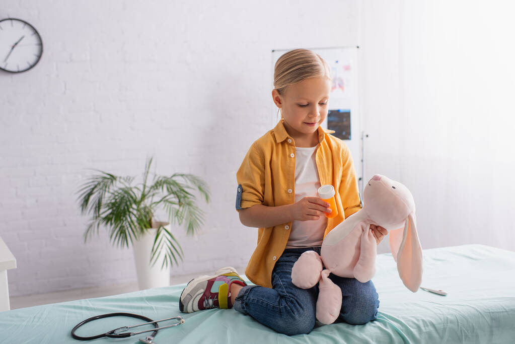 Meisje met pillen in de buurt van zacht speelgoed en stethoscoop op medische bank  - Foto, afbeelding