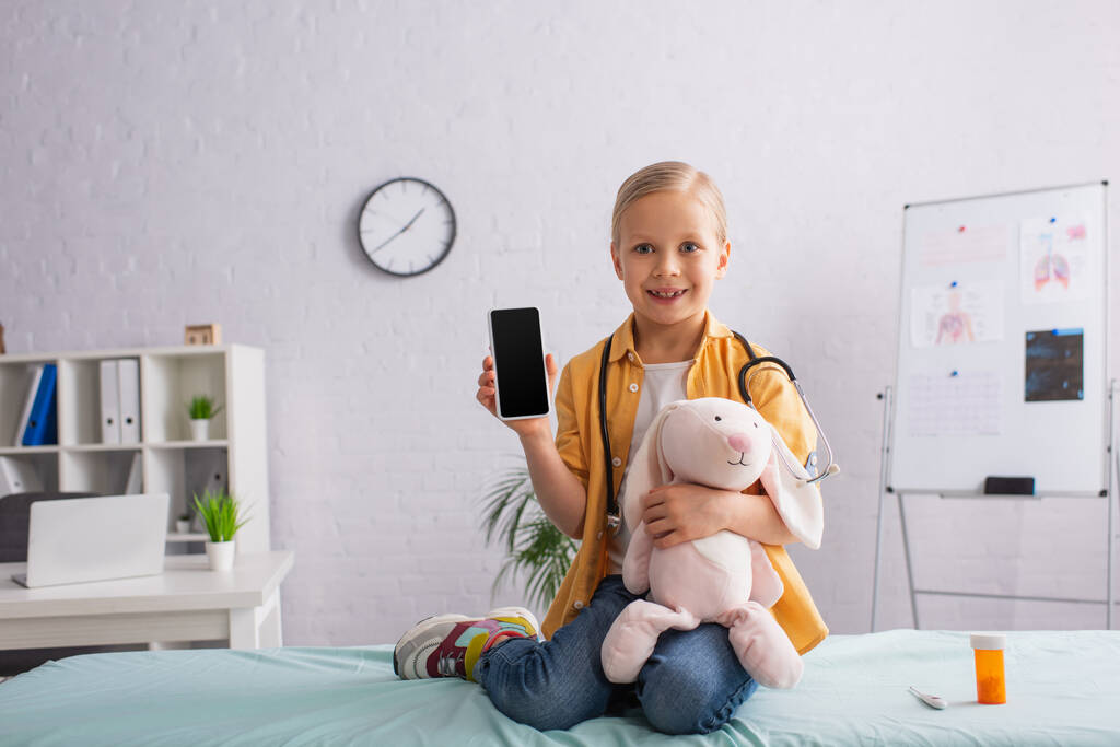 счастливая девушка сидит на медицинском диване с игрушечным кроликом и показывает смартфон с пустым экраном - Фото, изображение