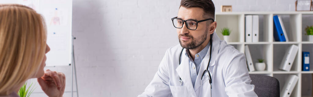 médico em casaco branco e óculos perto de paciente loira na clínica, banner - Foto, Imagem