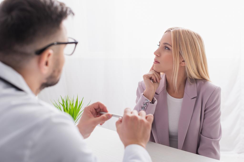 blonde woman thinking near blurred doctor during appointment in clinic - Photo, Image