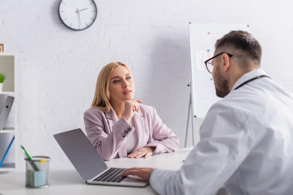 doctor and blonde woman talking in consulting room near laptop with blank screen - Photo, Image