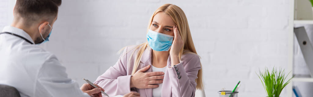 άρρωστη γυναίκα με ιατρική μάσκα που αγγίζει το κεφάλι και το στήθος κοντά σε θολή γιατρό, πανό - Φωτογραφία, εικόνα
