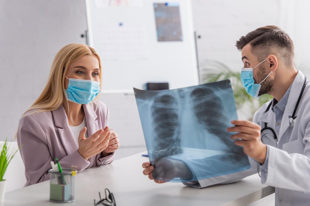 γιατρός με ιατρική μάσκα δείχνει πνεύμονες ακτινογραφία σε θετική γυναίκα κατά τη διάρκεια της διαβούλευσης - Φωτογραφία, εικόνα