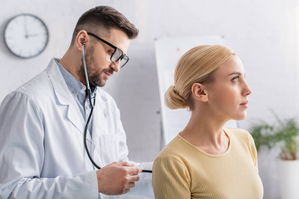 medico in cappotto bianco e occhiali esaminando donna bionda con stetoscopio - Foto, immagini