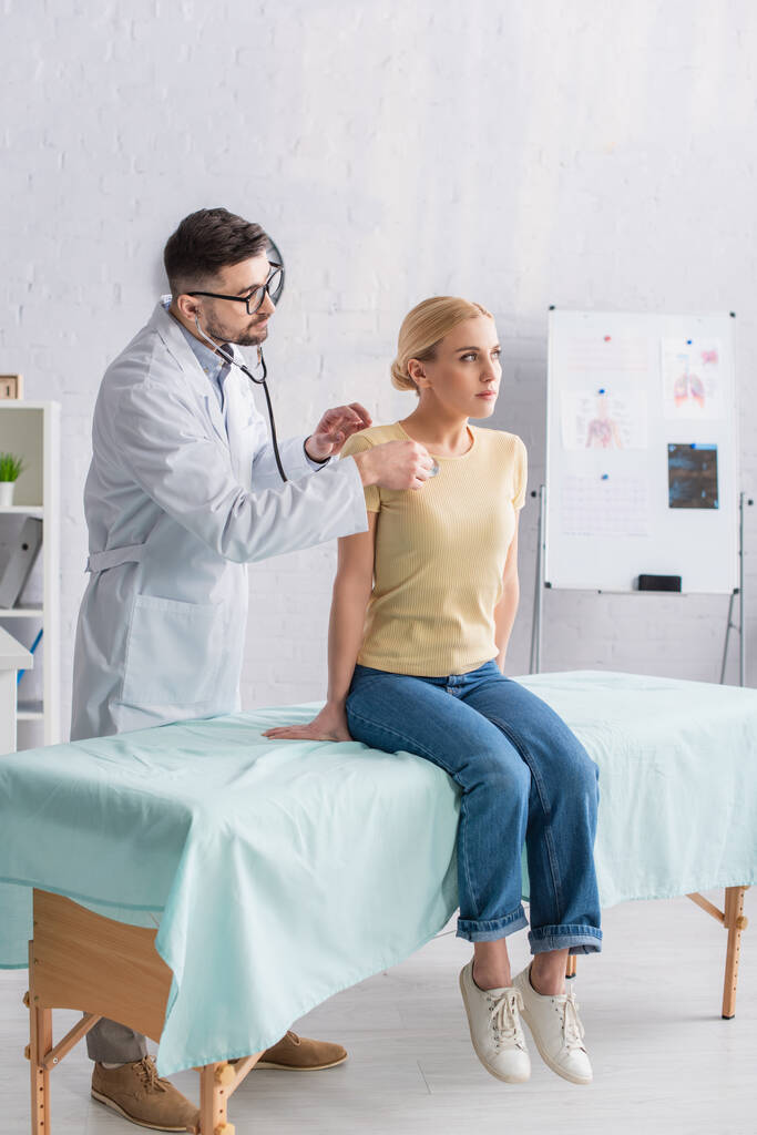 donna in jeans seduta sul divano medico mentre il medico la esamina con stetoscopio - Foto, immagini