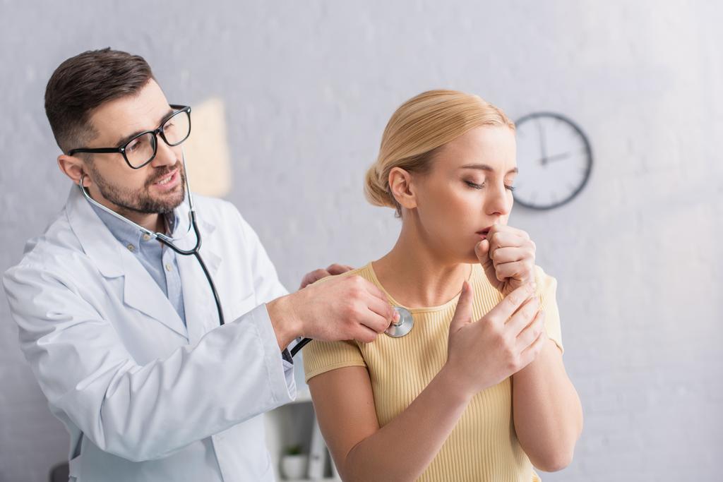 zieke vrouw hoesten in de buurt arts onderzoek haar met stethoscoop in kliniek - Foto, afbeelding