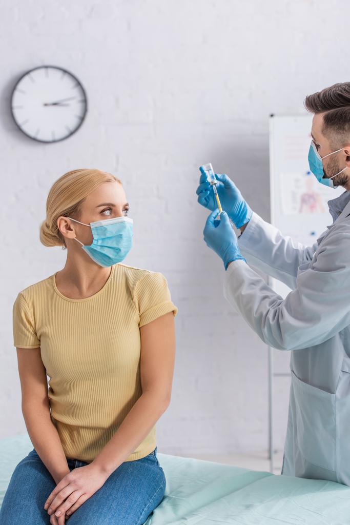 医者が注射器をワクチンで満たすのを見る保護マスクの女性 - 写真・画像