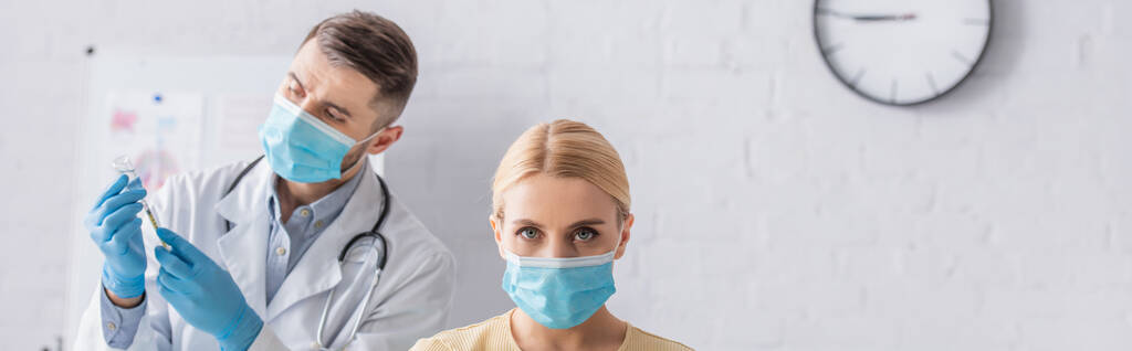 orvosi maszkos nő, aki a kamerába néz, miközben az orvos oltóanyaggal tölti meg a fecskendőt, banner - Fotó, kép