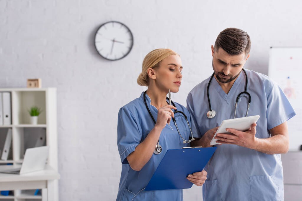médicos reflexivos en uniforme azul mirando tableta digital en el hospital - Foto, Imagen