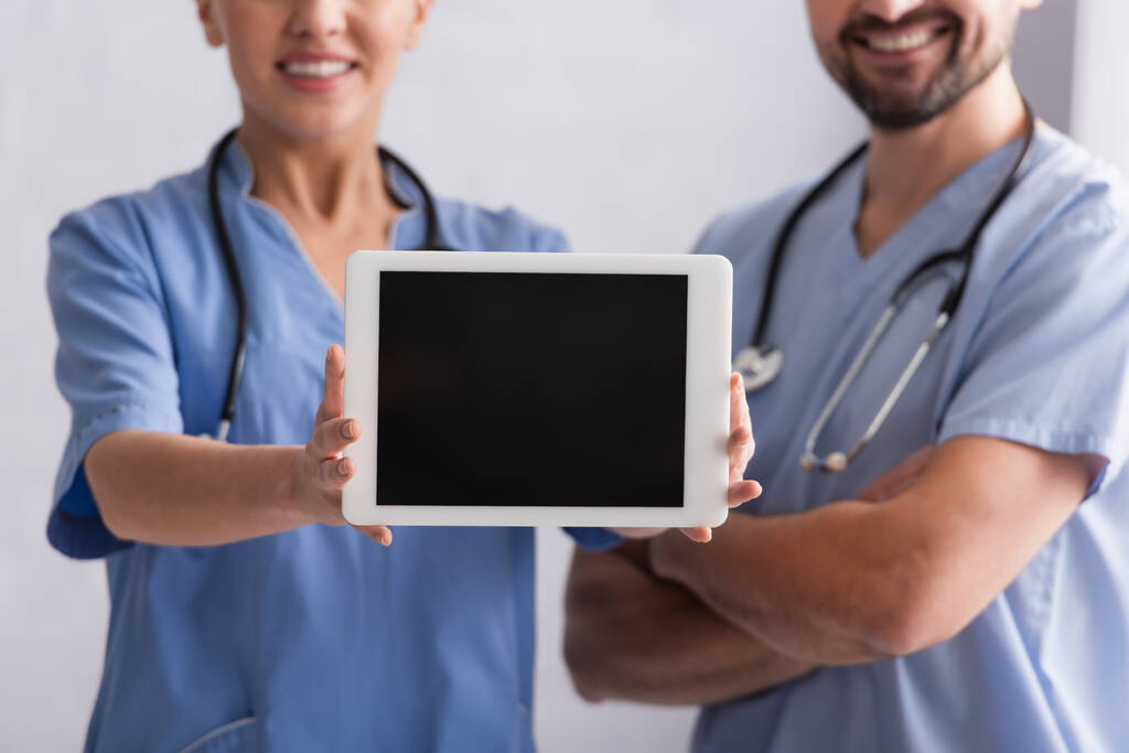 przycięty widok uśmiechniętego lekarza pokazującego cyfrową tabletkę w pobliżu kolegi z skrzyżowanymi ramionami - Zdjęcie, obraz