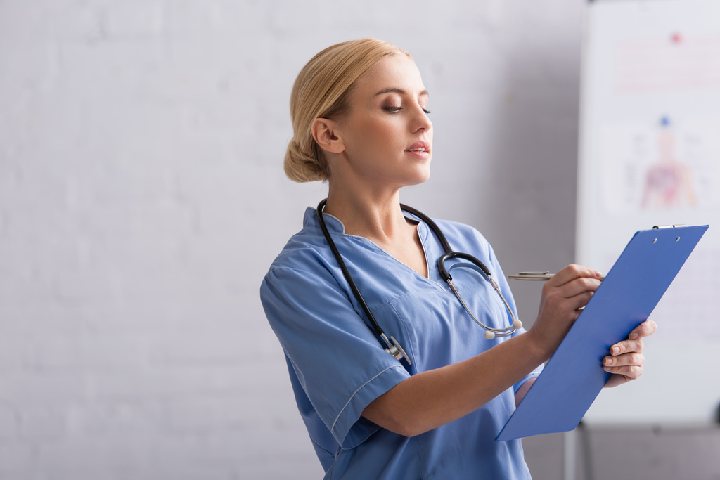 médecin en uniforme bleu écriture diagnostic sur presse-papiers en clinique - Photo, image
