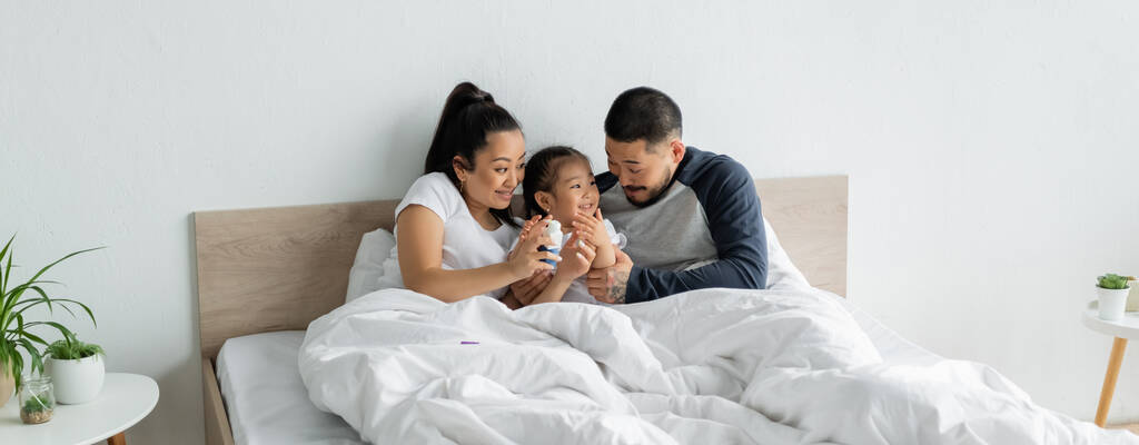 aasialainen äiti tilalla rakennuspalikoita lähellä iloinen lapsi ja aviomies sängyssä, banneri - Valokuva, kuva