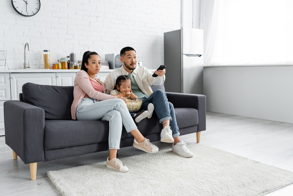 focused asian family watching movie and kid eating popcorn in living room - Photo, Image