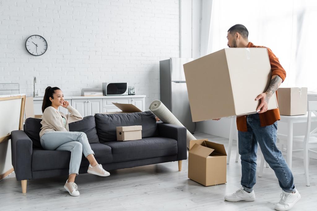 asian man holding carton box and looking at wife sitting on couch - Photo, Image