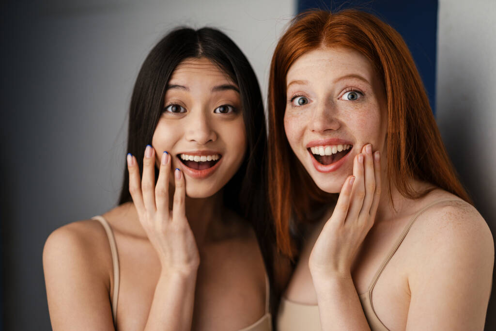 Gros plan portrait studio de deux belles jeunes filles excitées avec claire peau fraîche jeune visage posant sur un mur blanc, regardant la caméra - Photo, image