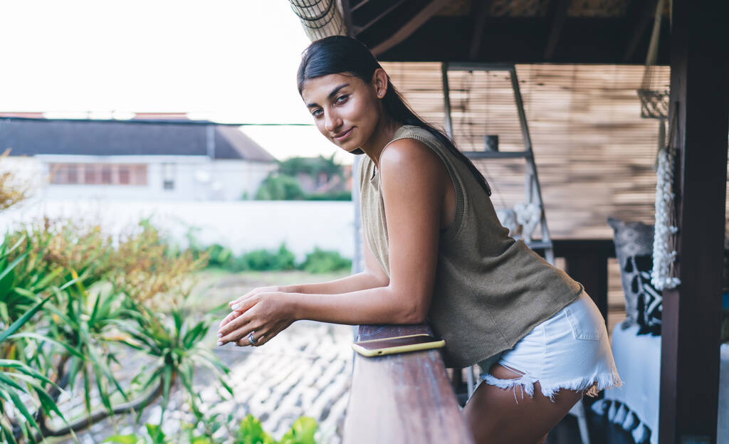 Vista lateral del contenido étnico femenino en traje de verano informal de pie en el balcón soleado en Bali resort y mirando a la cámara - Foto, imagen