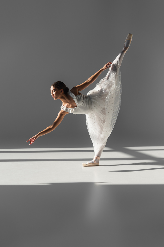 Vista laterale della ballerina in piedi su una gamba su sfondo grigio  - Foto, immagini