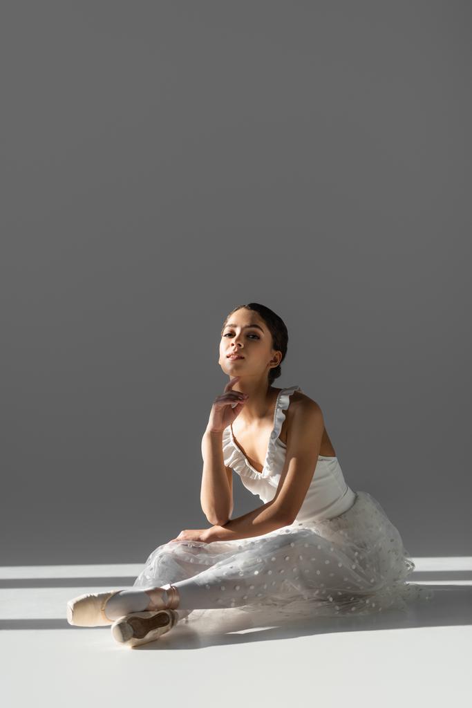 Brunette ballerina kijkend naar camera op grijze achtergrond met zonlicht - Foto, afbeelding