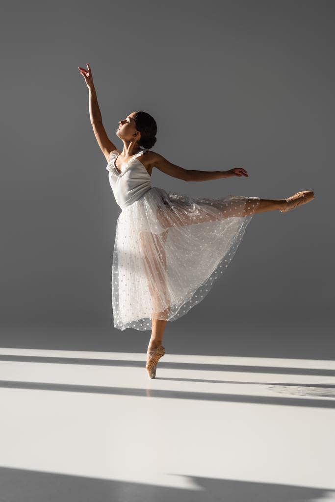 Zijaanzicht van elegante ballerina dansen op grijze achtergrond met zonlicht - Foto, afbeelding