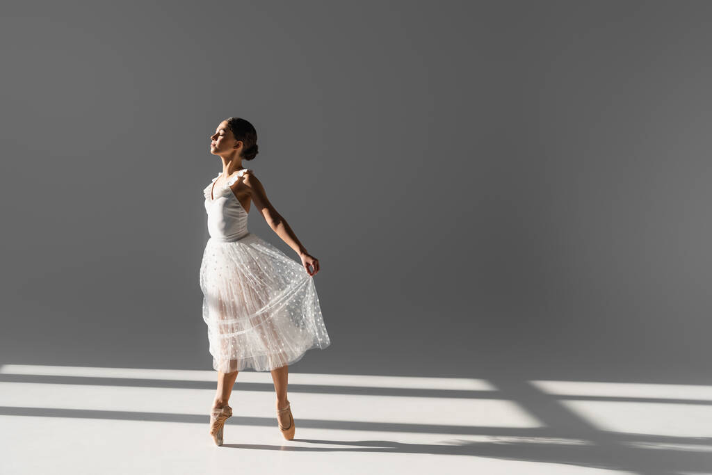 Вид сбоку на Фелину, держащую юбку во время танца на сером фоне с солнечным светом - Фото, изображение