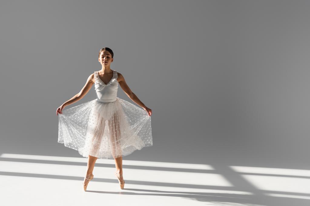 Longueur totale de ballerine en jupe et pointes dansant sur fond gris avec lumière du soleil - Photo, image