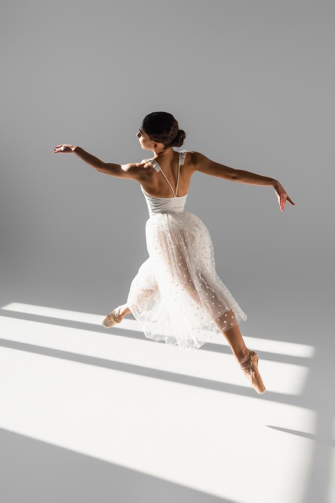 Vista laterale di elegante ballerina che salta alla luce del sole su sfondo grigio  - Foto, immagini