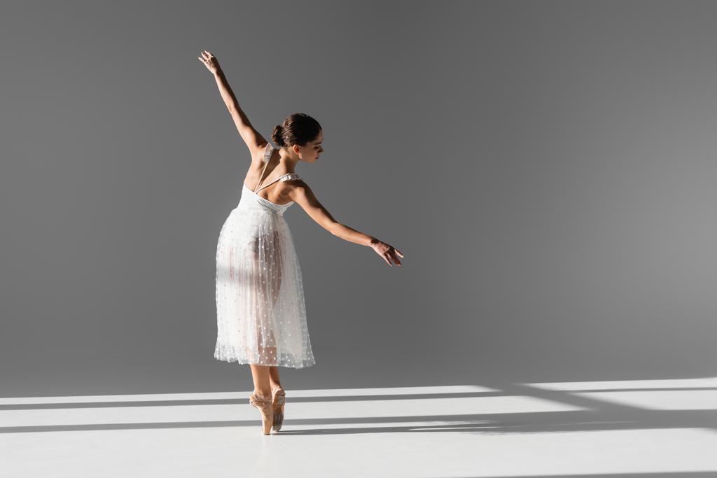 Zijaanzicht van elegante ballerina in puntschoenen op grijze achtergrond met zonlicht - Foto, afbeelding