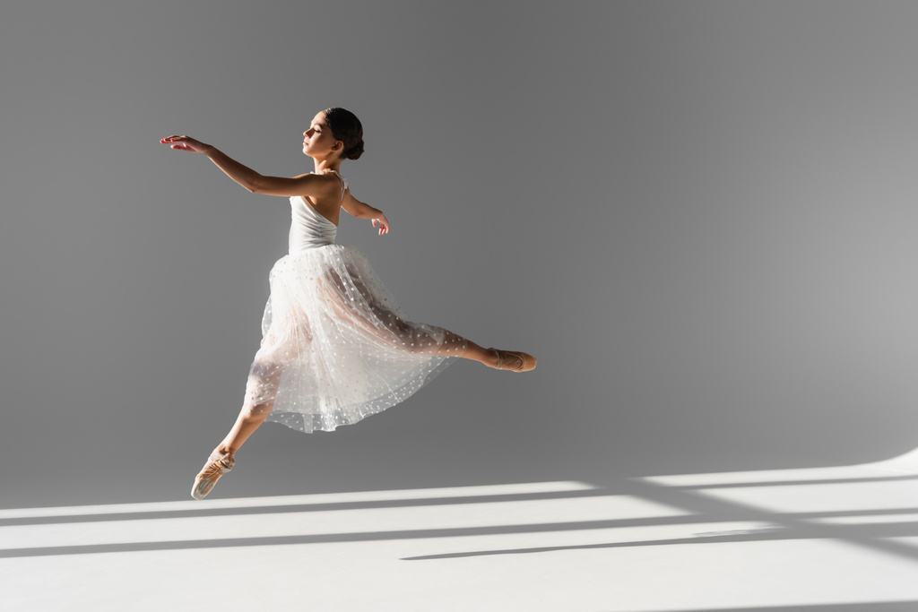 Gri arka planda güneş ışığıyla zıplayan profesyonel balerinin yan görüntüsü - Fotoğraf, Görsel