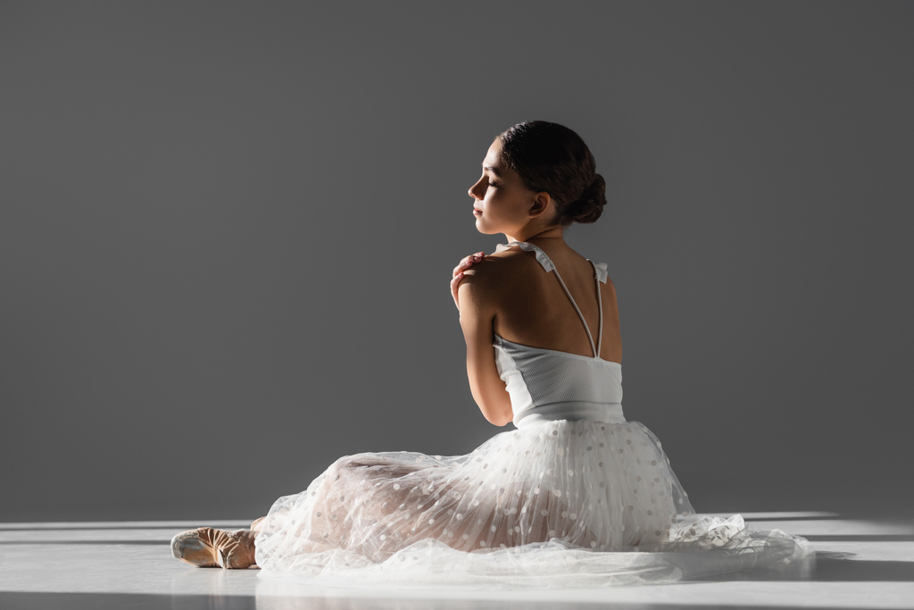 Boční pohled na mladou baletku dotýkající se ramene na šedém pozadí se slunečním světlem - Fotografie, Obrázek