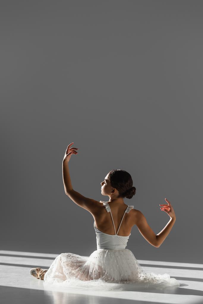 Vista lateral de la bailarina en vestido blanco y zapato puntiagudo bailando en suelo sobre fondo gris con luz solar - Foto, Imagen