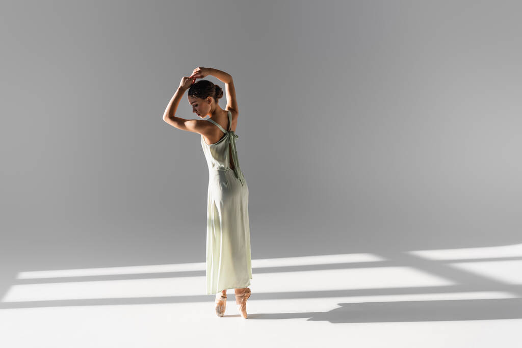 Vista laterale della ballerina bruna in scarpe da punta e vestito su sfondo grigio con luce solare - Foto, immagini