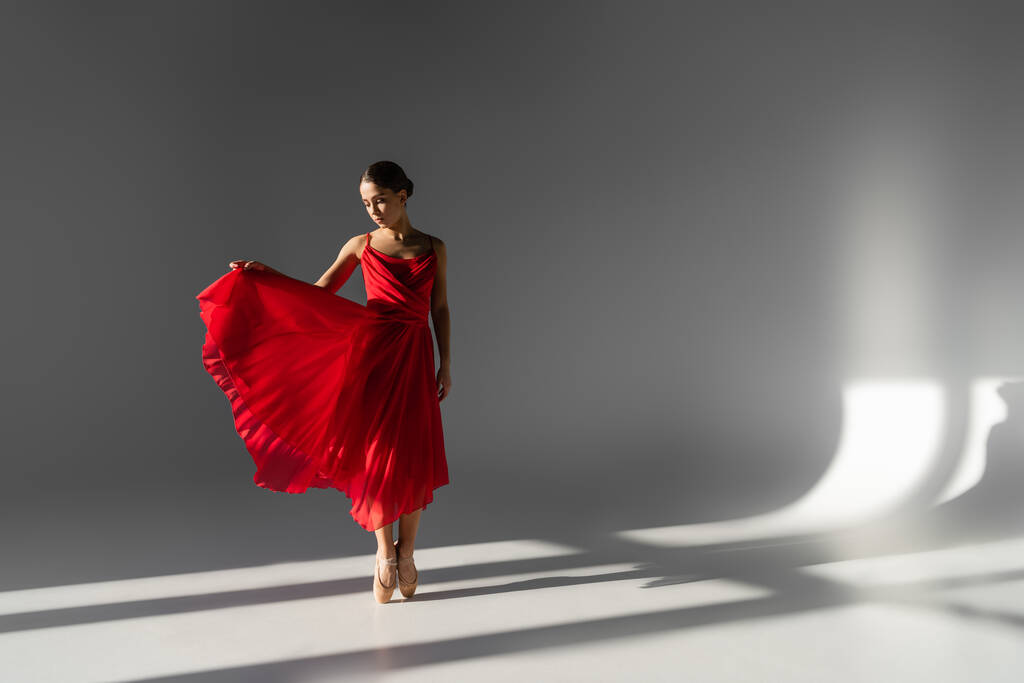 Bailarina en zapatos puntiagudos sosteniendo vestido rojo sobre fondo gris con luz solar - Foto, Imagen