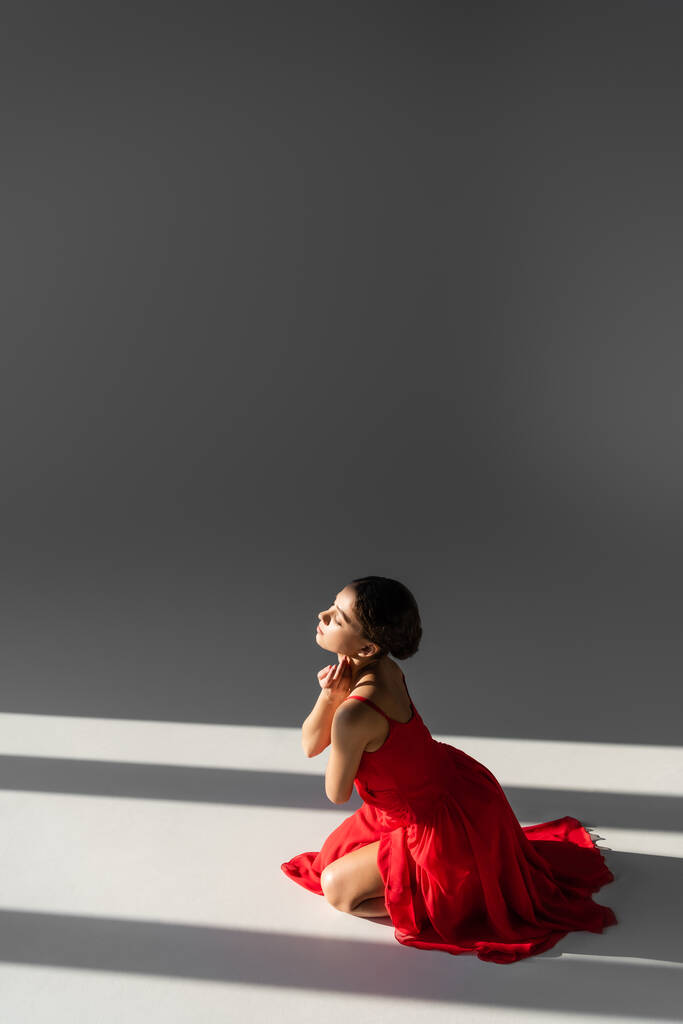 Zijaanzicht van brunette danser in rode jurk zitten op grijze achtergrond met zonlicht - Foto, afbeelding