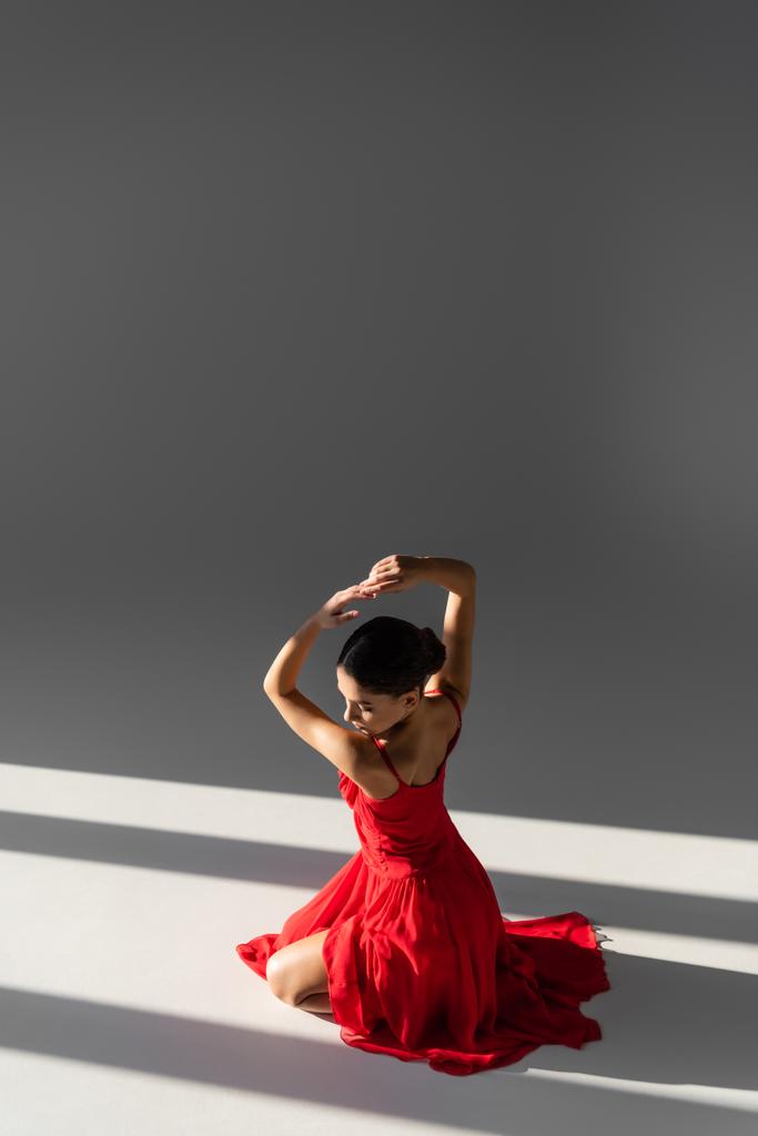 Gri arkaplanda güneş ışığıyla poz veren kırmızı elbiseli güzel bir dansçı. - Fotoğraf, Görsel