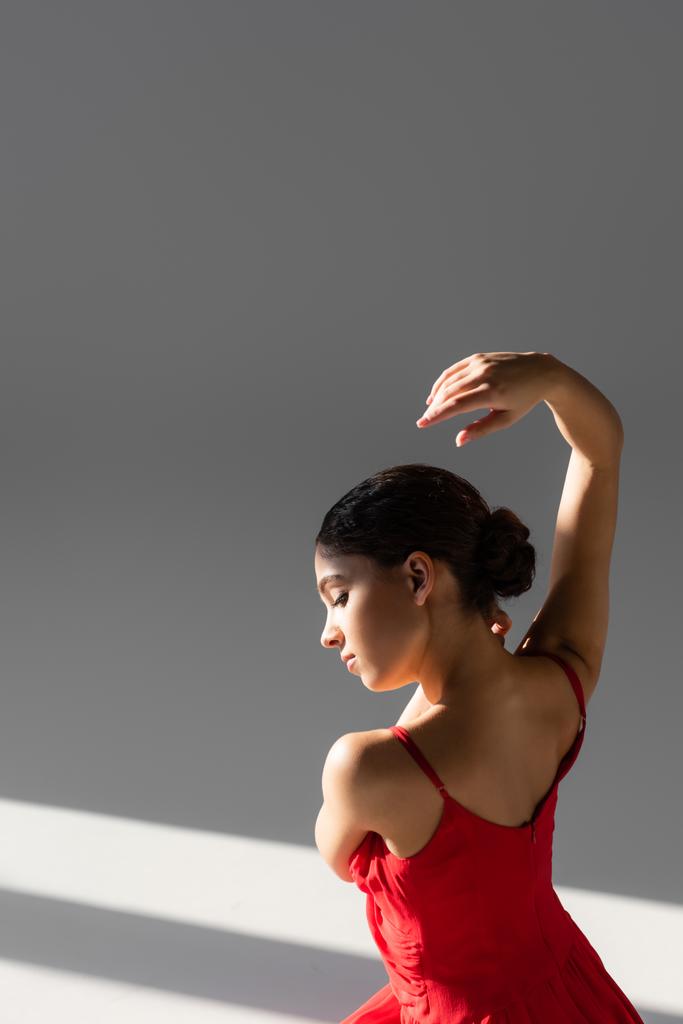 Boční pohled na klasickou tanečnici v červených šatech na šedém pozadí se slunečním světlem - Fotografie, Obrázek