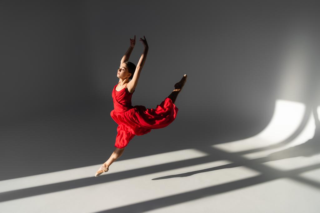 Вид збоку красивої балерини в червоній сукні, стрибки і підняття рук на сірому фоні з сонячним світлом
 - Фото, зображення