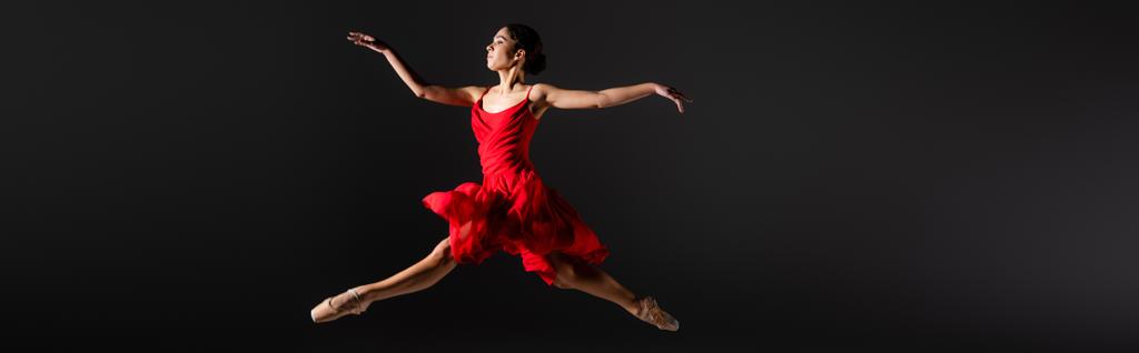 Side view of ballerina in pointe shoes and red dress jumping isolated on black, banner  - Photo, Image