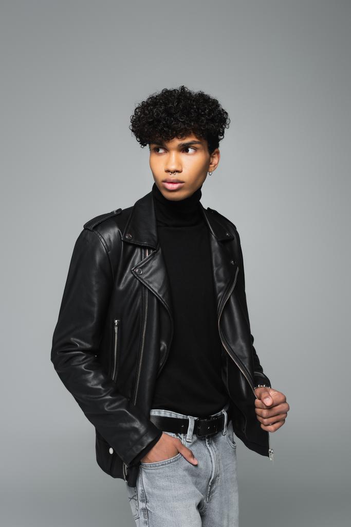 african american man in black leather jacket looking away while standing with hand in pocket of jeans isolated on grey - Foto, Imagem
