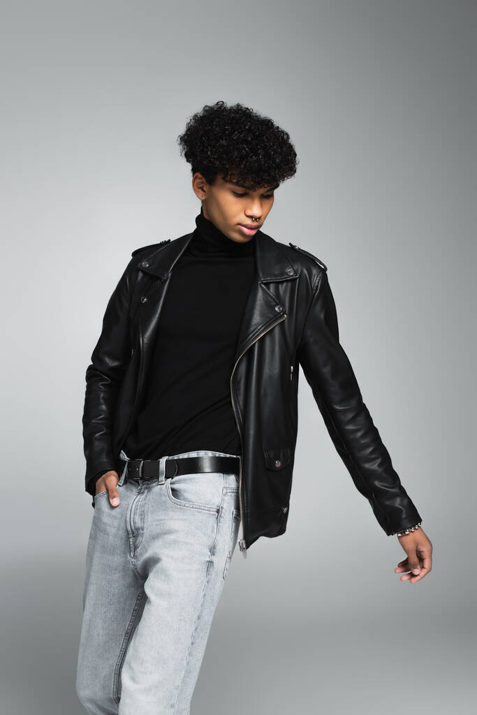 african american man with trendy hairstyle, in leather jacket, posing with hand in pocket on grey  - Foto, Imagem