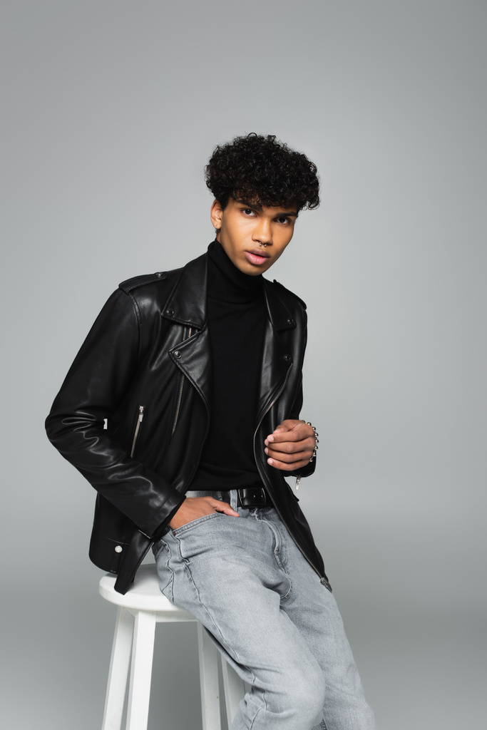 african american man in jeans and leather jacket sitting on stool while holding hand in pocket isolated on grey - Photo, image