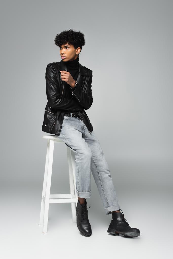 молодий афроамериканський чоловік у шкіряній куртці та джинсах дивиться на високий табурет на сірому фоні
 - Фото, зображення